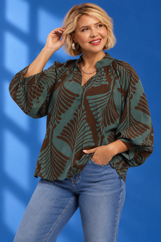 A woman wearing a green and brown patterned blouse with three-quarter sleeves and blue jeans, standing against a blue studio backdrop.