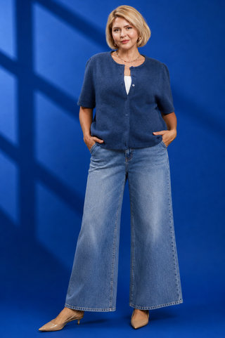 A woman modeling a short-sleeve blue knit cardigan over a white top, paired with wide-leg blue jeans and beige heels, standing against a blue studio backdrop with window shadow patterns.