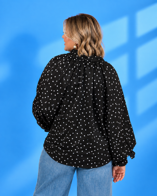 A woman shown from the back wearing a black long-sleeve blouse with white polka dots and light blue jeans