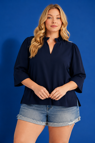 A woman modeling a navy blue blouse with a split V-neckline and three-quarter bell sleeves, paired with denim shorts, against a solid blue studio background.