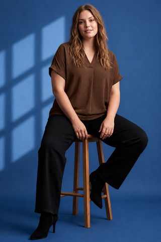 A woman sits on a wooden stool wearing a brown short-sleeve V-neck top and black wide-leg pants against a blue studio backdrop with window shadow projections.
