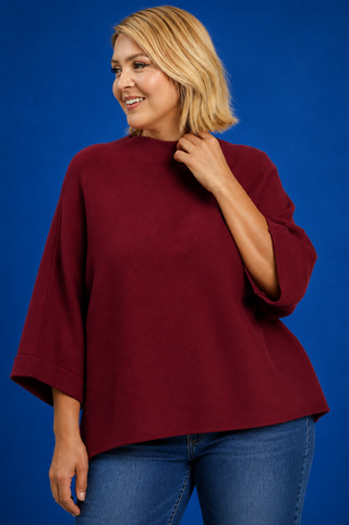 A woman with blonde hair wearing a loose-fitting, high-neck burgundy top and blue jeans, posing against a solid blue studio background.