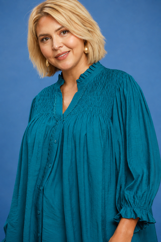 A woman with blonde hair wearing a teal blue smocked blouse with ruffled trim, photographed against a solid blue studio background.