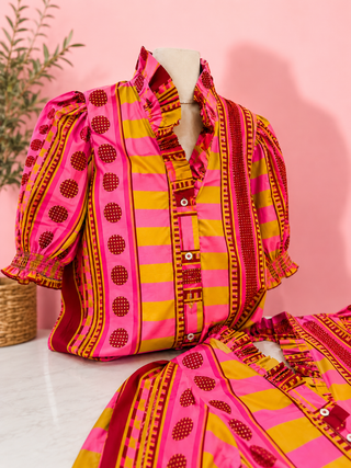 A vibrant pink and yellow patterned blouse displayed on a mannequin torso, with a matching skirt or shorts partially visible in the foreground. The blouse features bold vertical stripes, puff sleeves, and red circular dot motif