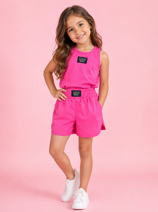 A young girl stands outdoors on a sunlit walkway wearing a light lavender sleeveless tank top and matching shorts