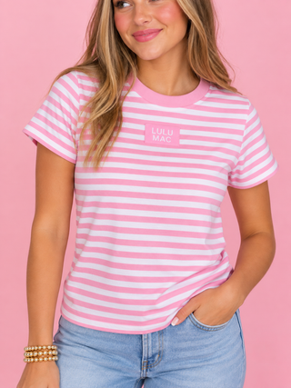 A woman wearing a pink and white striped short-sleeve t-shirt with a pink chest logo that reads 'LULU MAC', paired with light blue jeans, set against a solid pink background.