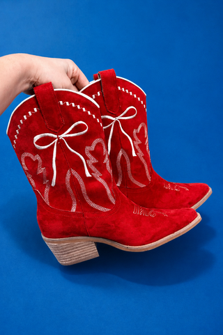 A pair of red suede cowboy boots with white embroidered bow and scroll designs, held by a hand against a solid blue backgrou