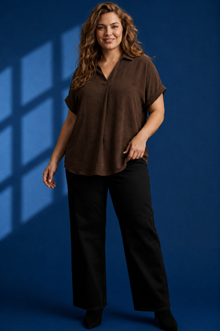 A woman modeling a brown short-sleeve button-up blouse with black wide-leg pants, standing against a dark blue studio backdrop with window shadow effects.