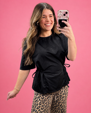 Woman taking a mirror selfie wearing a black top and leopard print pants against a pink background
