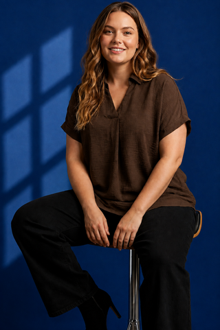 A woman with long wavy brown hair sits on a stool against a dark blue background, wearing a brown short-sleeve collared blouse and black pants.