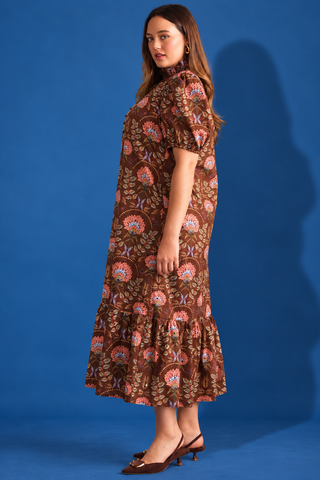 A woman modeling a brown midi dress with a colorful floral and paisley print, standing against a solid blue studio background.