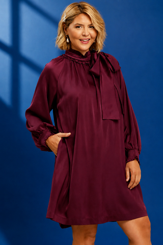 A woman modeling a long-sleeve, high-neck, boat-neck dress in deep burgundy with a large bow at the collar, set against a dark blue studio background.