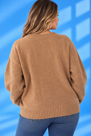 A woman shown from the back wearing a camel brown knit sweater with textured eyelet details on the sleeves, paired with blue jeans