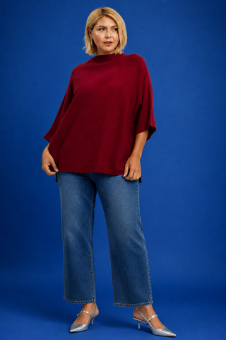 A woman modeling a loose-fitting, deep burgundy mock-neck top with wide three-quarter sleeves, paired with blue wide-leg jeans and silver heels, set against a solid blue studio background.