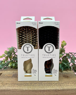 Two Teleties boar bristle brush packages on a wooden surface with a pink background and greenery.