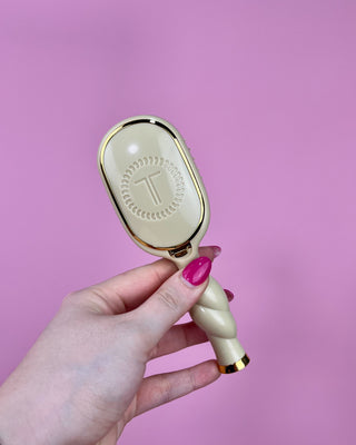 Hand holding a white and gold hairbrush against a pink background