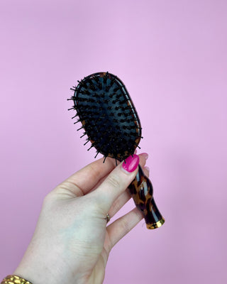 Hand holding a hairbrush with a tortoiseshell handle against a pink background
