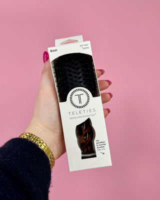 Hand holding a packaging of Teleties hairbrush against a pink background