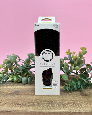 Teleties hairbrush packaging on a wooden surface with greenery and a pink background