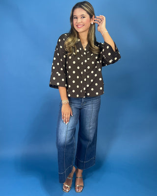 A woman modeling a dark brown button-up blouse with white polka dots and wide three-quarter sleeves, paired with blue jea