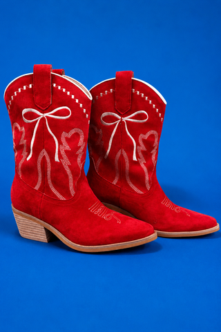 A pair of red suede cowboy boots with white western embroidery, including bow and heart designs, set against a solid blue ba