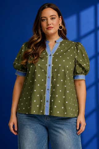 A woman modeling a short-sleeve button-front blouse with puff sleeves, olive green fabric, white polka dots, and light blue trim, paired with blue jeans against a blue studio backdrop.