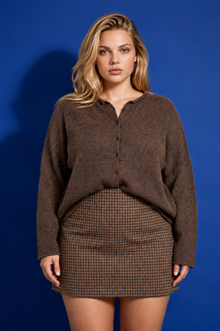 A woman modeling a brown fuzzy long-sleeve button-front top and a brown-and-black checkered mini skirt against a solid blue studio background.