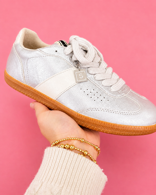 White sneaker with brown sole held by a hand against a pink background