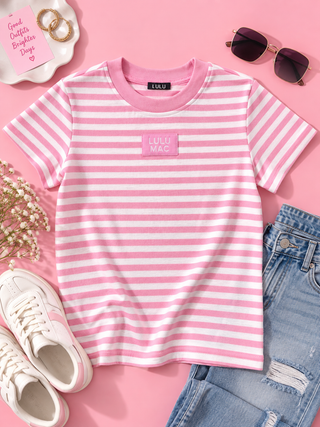 Pink and white striped short-sleeve t-shirt with a pink chest patch reading 'LULU MAC', styled in a flat lay with blue jeans, white sneakers, sunglasses, and a pink card on a pink background.