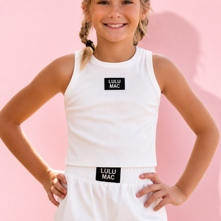 A young girl stands outdoors on a sunlit walkway wearing a light lavender sleeveless tank top and matching shorts