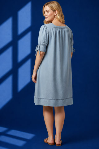 A woman wearing a light blue puff-sleeve dress, shown from the back against a dark blue studio backdrop with window shadow patterns.