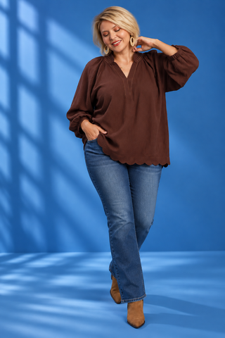A woman modeling a brown long-sleeve blouse with a scalloped hem and blue jeans, standing against a blue studio backdrop with window shadow patterns.
