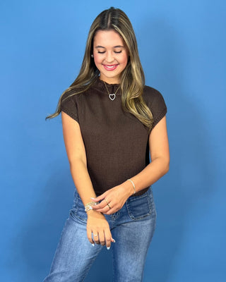 a brown short-sleeve ribbed knit top and blue jeans, standing against a solid blue background.