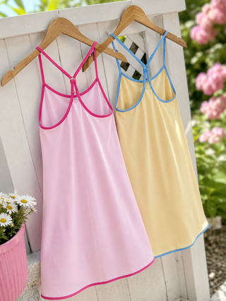 Two sleeveless babydoll-style camisole dresses hanging on wooden hangers against a white wooden fence, with pink trim on the