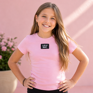 wearing a light pink crewneck t-shirt with a black rectangular patch on the chest that reads 'LULU MAC' in white.