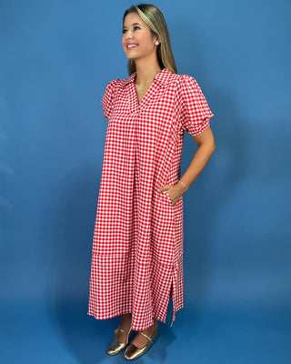 A woman modeling a red and white gingham wrap-style midi dress with short puff sleeves, standing against a solid blue studio