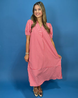 A woman modeling a red and white gingham wrap-style midi dress with short puff sleeves, standing against a solid blue studio