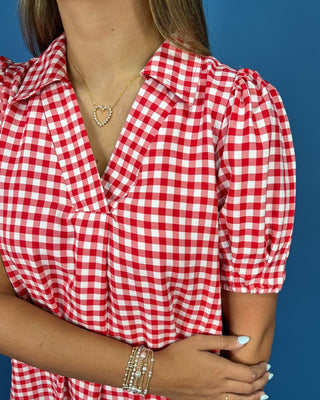A close-up of a woman wearing a red and white gingham short-sleeve blouse with puff sleeves, paired with a heart-shaped neckl