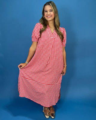 A woman modeling a red and white gingham wrap-style midi dress with short puff sleeves, standing against a solid blue studio