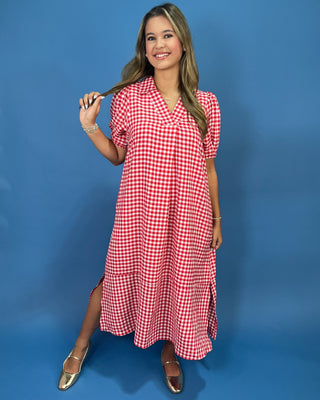 A woman modeling a red and white gingham wrap-style midi dress with short puff sleeves, standing against a solid blue studio