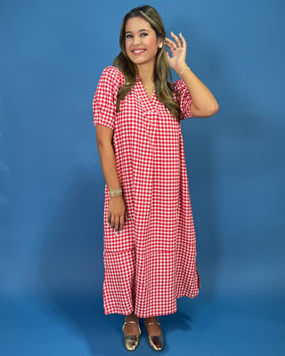 A woman modeling a red and white gingham midi dress with short sleeves and a V-neckline, standing against a solid blue studi