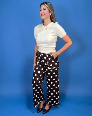A woman modeling a white short-sleeve knit top and black wide-leg pants with white polka dots, standing aga