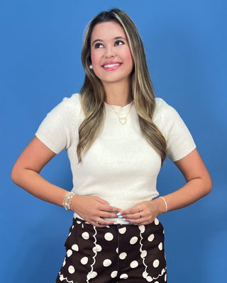 A woman posing against a solid blue background, wearing a cream short-sleeve knit top and dark brown polka-dot skirt with wh