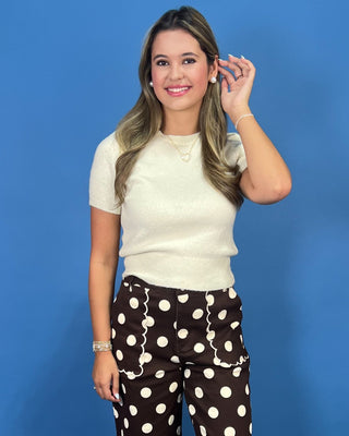 A woman wearing a short-sleeve cream knit sweater and brown polka-dot pants, standing against a solid blue background.