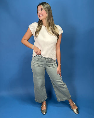 A woman posing in a cream ribbed short-sleeve crop top and gray jeans against a solid blue studio background.