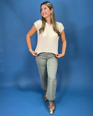 A woman posing in a cream ribbed short-sleeve crop top and gray jeans against a solid blue studio background.