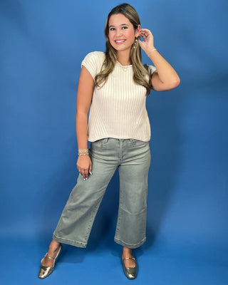 A woman posing in a cream ribbed short-sleeve crop top and gray jeans against a solid blue studio background.