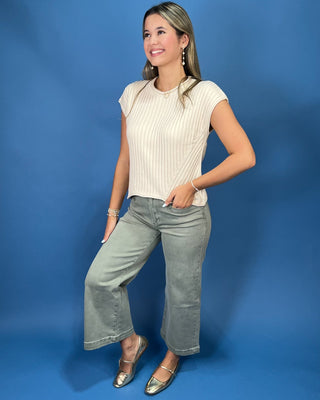 A woman posing in a cream ribbed short-sleeve crop top and gray jeans against a solid blue studio background.