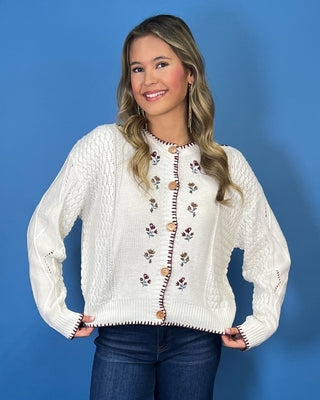 a cream cable-knit button-up sweater with embroidered floral details and brown buttons