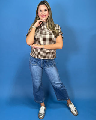 A woman posing in a brown and beige checkered short-sleeve mock neck top, blue cropped jeans, and white sneakers against a s